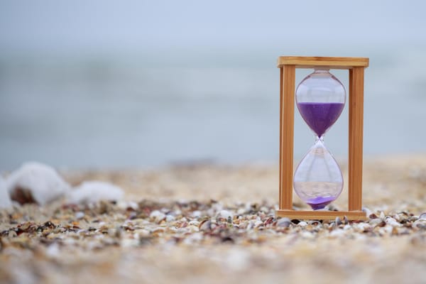 An hourglass with purple sand standing on a beach, with the ocean blurred in the background.